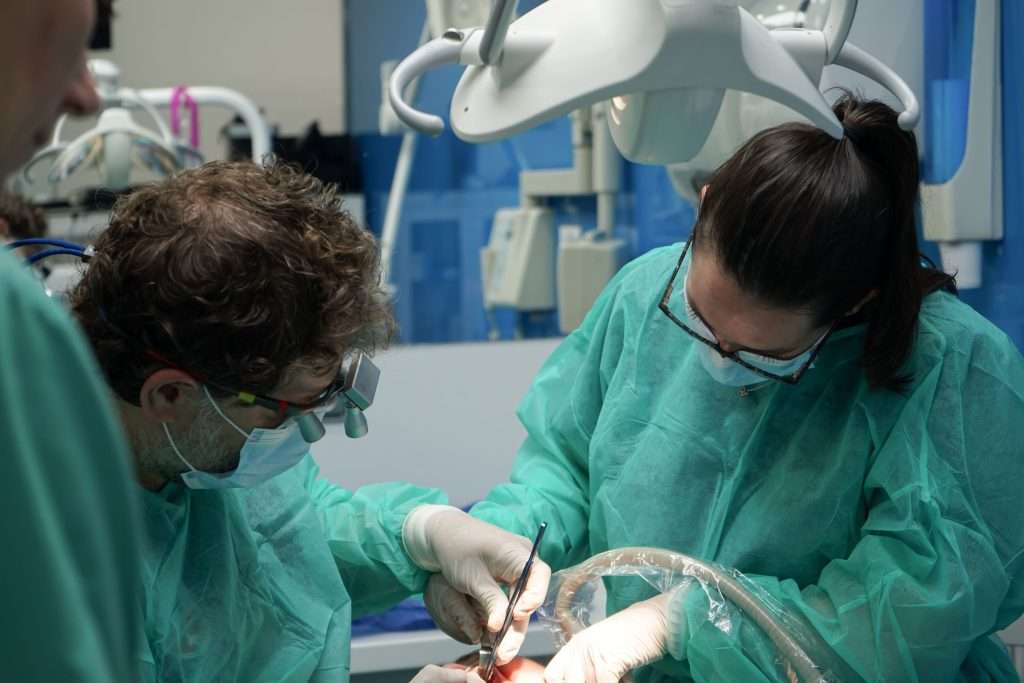 Dos cirujanos con batas verdes y mascarillas operan bajo luces brillantes en una moderna clínica dental de Moratalaz. de Hospital Dental de Moratalaz "Martínez y Morcuende"