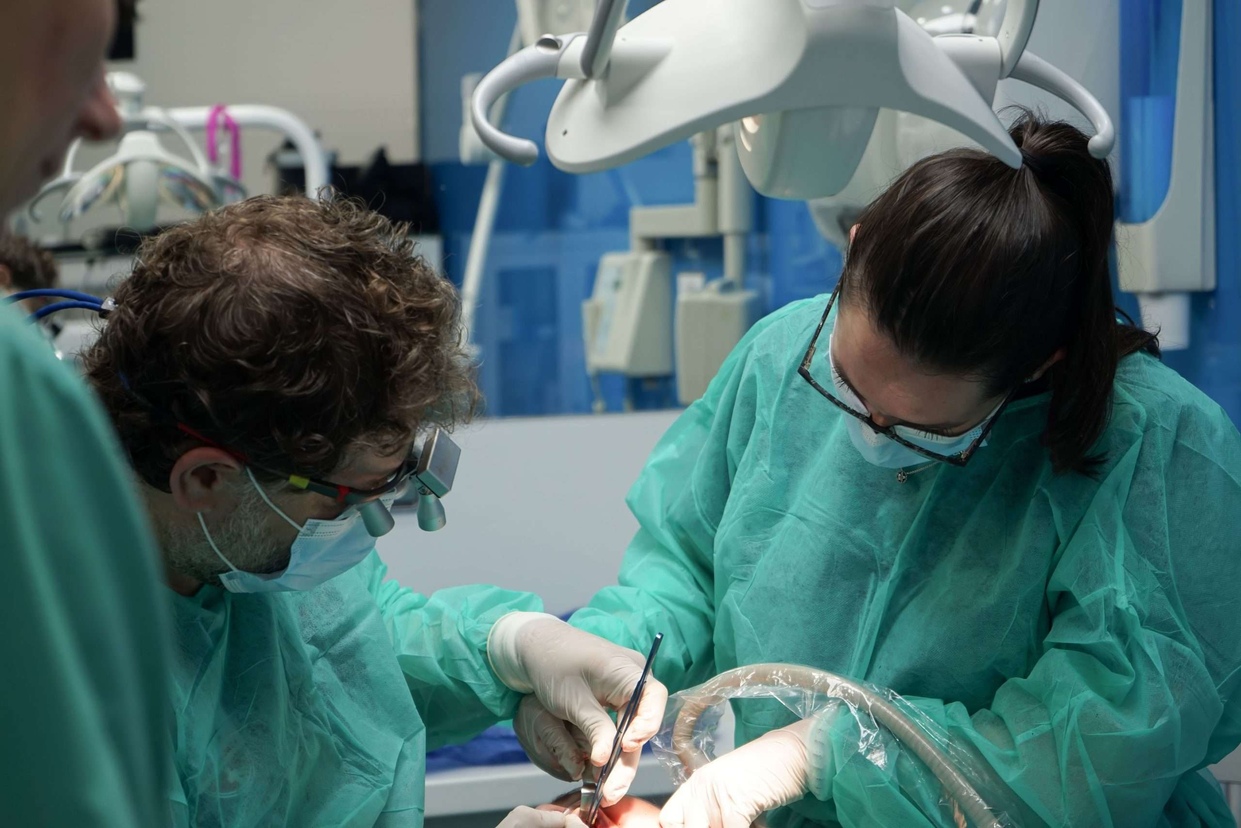 Dos cirujanos con batas verdes y mascarillas operan bajo luces brillantes en una moderna clínica dental de Moratalaz. de Hospital Dental de Moratalaz "Martínez y Morcuende"