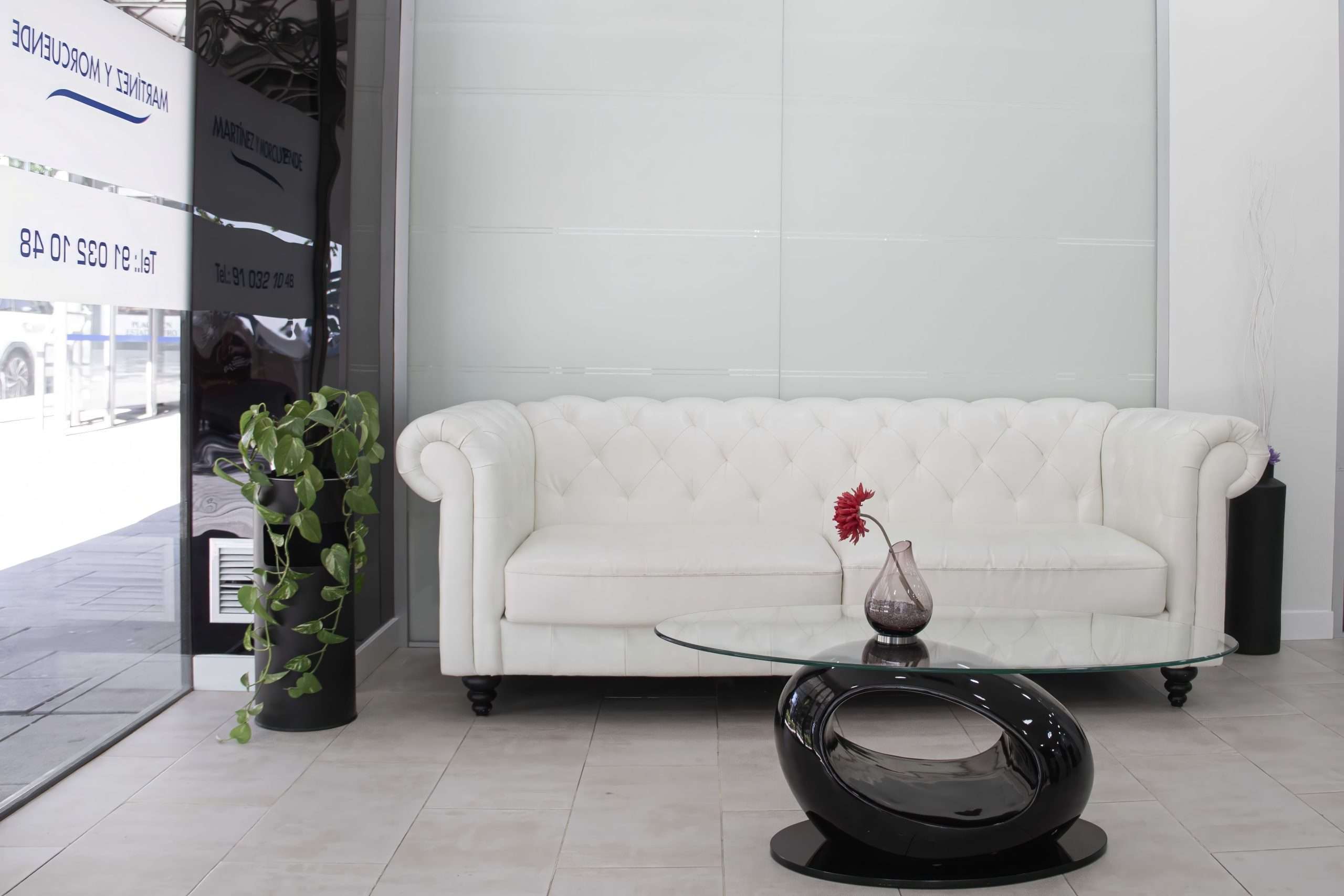 Una moderna sala de espera dental en Moratalaz con un sofá blanco empenachado, una mesa de centro de cristal, un jarrón de flores rojas y una planta en maceta. de Hospital Dental de Moratalaz "Martínez y Morcuende"
