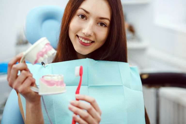 Mujer en un sillón dental en una clínica dental Moratalaz, sosteniendo un cepillo de dientes y un modelo, sonriendo a la cámara con un babero dental azul. de Hospital Dental de Moratalaz "Martínez y Morcuende"