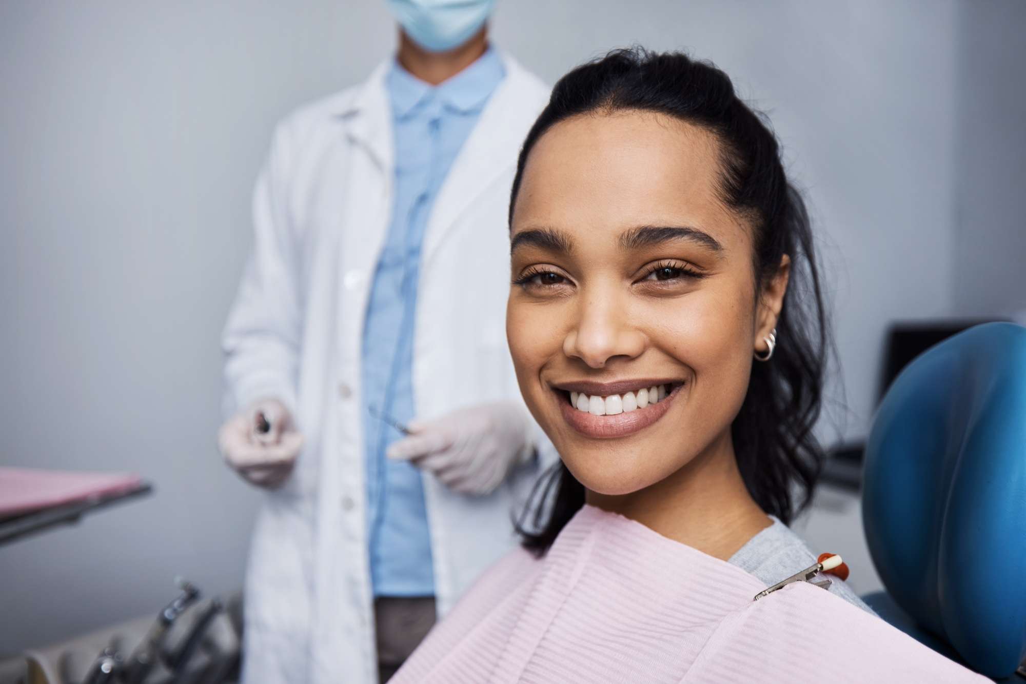 Mujer sonriente sentada en el sillón de una clínica dental con un odontólogo con bata blanca y guantes de pie al fondo. de Hospital Dental de Moratalaz "Martínez y Morcuende"