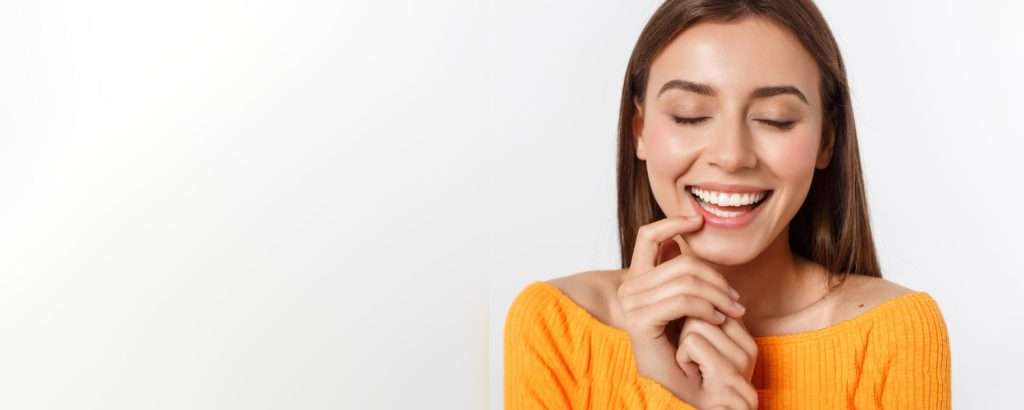 Una joven con un top naranja brillante sonríe con los ojos cerrados, mostrando su salud dental sobre un fondo blanco. de Hospital Dental de Moratalaz "Martínez y Morcuende"