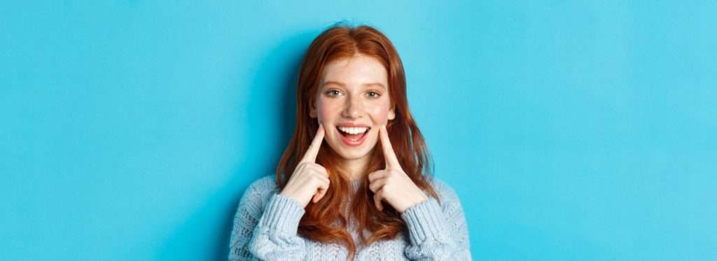 Una mujer sonriente de larga melena pelirroja se señala las mejillas, mostrando salud dental, de pie frente a un fondo azul. de Hospital Dental de Moratalaz "Martínez y Morcuende"