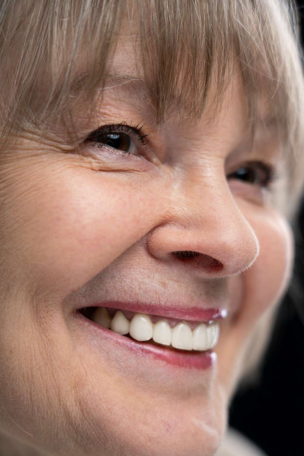 Primer plano de una mujer mayor sonriendo, mostrando los dientes, en el exterior de una clínica dental en Moratalaz. El pelo le cubre ligeramente la frente. de Hospital Dental de Moratalaz "Martínez y Morcuende"