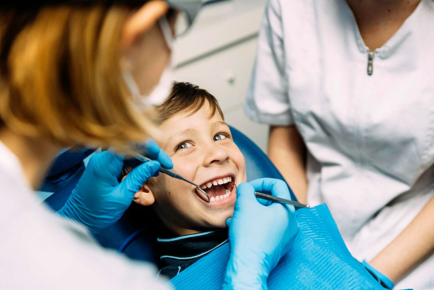 Un niño sonríe en una clínica dental de Moratalaz mientras un odontólogo le examina los dientes, otra persona a su lado. de Hospital Dental de Moratalaz "Martínez y Morcuende"