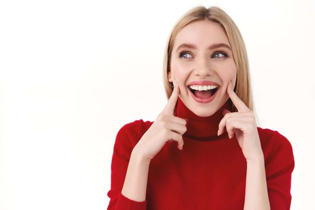 Mujer sonriente con jersey rojo se señala las mejillas, resaltando su salud dental sobre fondo blanco. de Hospital Dental de Moratalaz "Martínez y Morcuende"