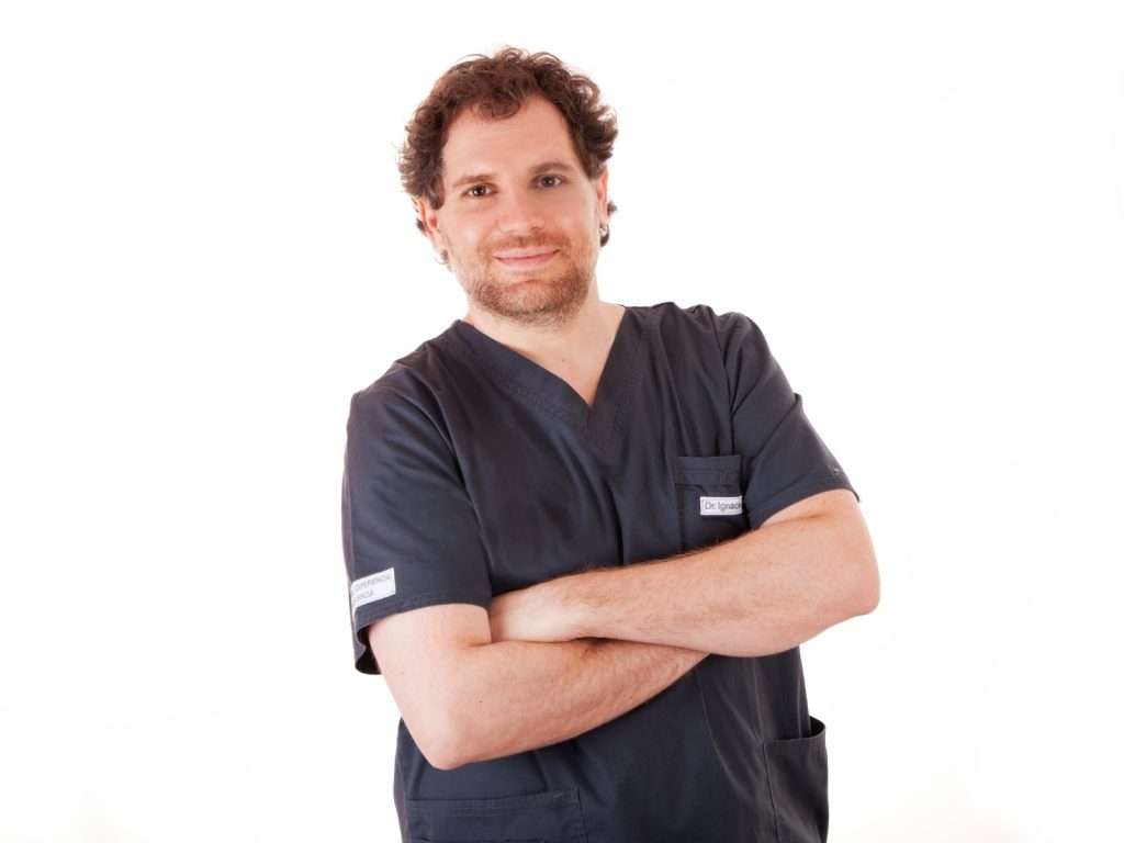 Un dentista con bata oscura sonríe con los brazos cruzados sobre un fondo blanco liso en una clínica dental. de Hospital Dental de Moratalaz "Martínez y Morcuende"